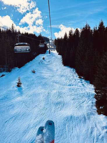A person rides a ski lift above a snowy slope, surrounded by dense evergreen trees. Blue sky, bright clouds, fresh ski tracks, and peaceful winter scenery.