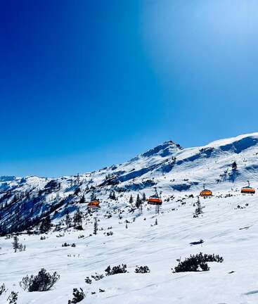 Bright blue sky over snowy mountain landscape, scattered pine trees, and orange ski lift cabins gliding uphill. Pristine winter scene, perfect for skiing and adventure.