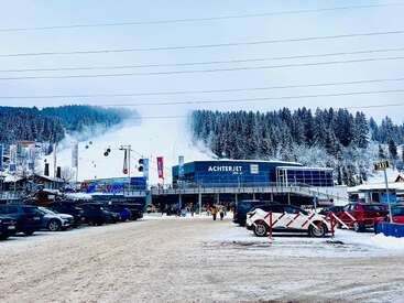 A snowy ski resort scene shows the "Achterjet" gondola station, parked cars, people entering, surrounding pine trees, and ski lifts ascending the white mountain slope.