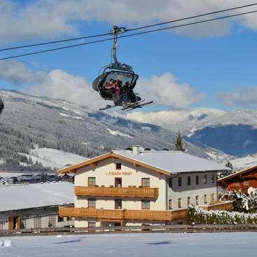 L'image montre une station de ski avec une remontée mécanique, avec un grand groupe de personnes, quelques arbres et un paysage montagneux. Le ciel est clair et ensoleillé. L'atmosphère générale est à la détente et aux loisirs.