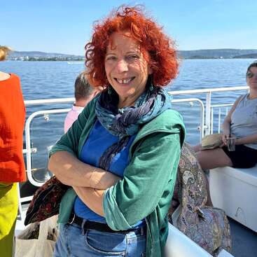 Eine lächelnde Frau mit lockigem rotem Haar steht auf einem Boot, die Arme verschränkt, und trägt ein blaues Hemd und eine grüne Jacke. Hinter ihr sieht man einen sonnigen Tag, Wasser und Menschen.