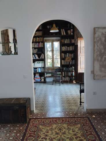 Ein gemütlicher Raum mit einem Torbogen führt in eine Bibliothek mit Bücherregalen, einem Fenster, einer antiken Truhe, gemusterten Fliesen, einem Orientteppich und klassischer Einrichtung.