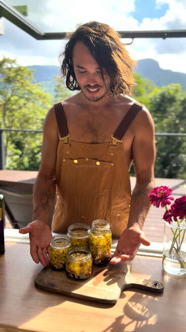 A man in brown overalls stands by a window, presenting four jars filled with pickled vegetables on a wooden board, with flowers in a vase nearby.