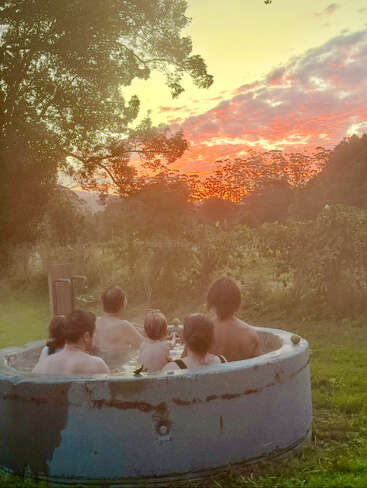 A group of people relaxes together in a round outdoor hot tub, surrounded by lush greenery, watching a colorful, peaceful sunset under a vibrant sky.