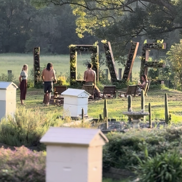 Four people gather near wooden chairs and a large “LOVE” sign made from plants in a lush, green garden with beehives and sunshine filtering through trees.