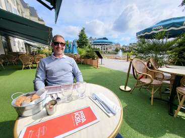 Um homem sorridente de óculos escuros senta-se em uma mesa de café ao ar livre com bolos e copos, cercado por vegetação, com vista para um rio panorâmico em um dia ensolarado.