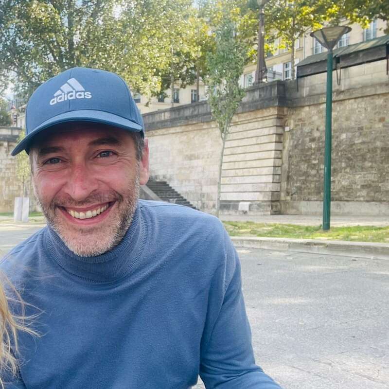 Um homem sorridente, usando um boné Adidas azul e um suéter azul, senta-se ao ar livre perto de um muro de pedra e degraus, com árvores e edifícios ao fundo.