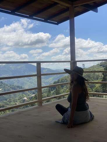 Uma mulher está sentada em um deck de madeira, olhando para uma paisagem montanhosa sob um céu azul com nuvens brancas. Ela usa um chapéu e uma camiseta regata.