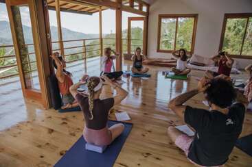 A imagem mostra um grupo de pessoas envolvidas em uma sessão de ioga ou meditação, sentadas no chão com as pernas cruzadas, cercadas por um ambiente sereno e pacífico.