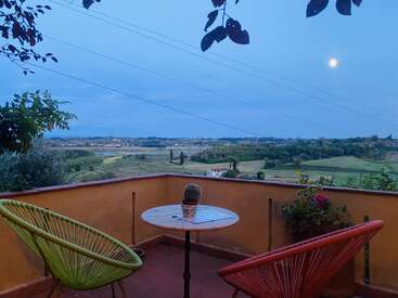 Um terraço tranquilo ao entardecer com duas cadeiras coloridas, um cacto em uma mesa redonda, com vista para campos verdes ondulados e uma lua cheia no céu.