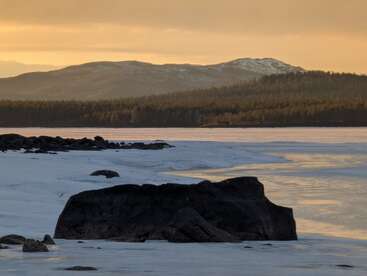 Un sereno paisaje invernal con la costa cubierta de nieve y hielo, rocas oscuras, aguas tranquilas, colinas boscosas y montañas lejanas bajo un cálido cielo dorado al atardecer.