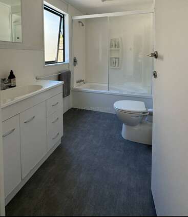Dieses Bad ist sauber, modern und minimalistisch. Es hat einen weißen Waschtisch, einen Spiegel, eine Toilette, eine Dusch-Badewannen-Kombination mit Glasschiebetüren und ein Fenster für natürliches Licht.