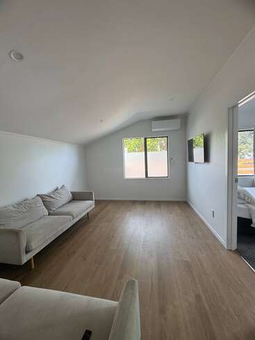 Das Bild zeigt ein minimalistisches Wohnzimmer mit hellem Holzboden, zwei beigen Sofas, einem an der Wand montierten Fernseher, einer Klimaanlage, einem Fenster und einem angrenzenden Schlafzimmer.