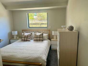Dieses gemütliche Schlafzimmer hat ein Doppelbett mit karierten Kissen, weiße Bettwäsche, zwei Nachttische mit Lampen, eine Kommode, ein Fenster und eine minimalistische Einrichtung.