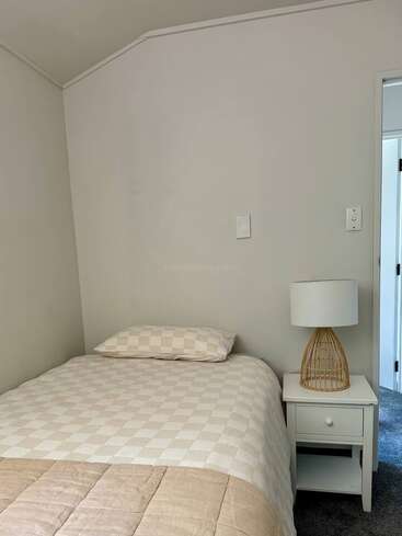 Das Bild zeigt ein einfaches Schlafzimmer mit einem Einzelbett, einer beige karierten Bettwäsche, einem passenden Kissen, einem weißen Nachttisch und einer Lampe mit einem geflochtenen Fuß.