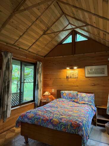 This cozy wooden cabin bedroom features a colorful sea-themed bedspread, soft lighting, large windows with nature views, sloped ceiling, rustic decor, and inviting, peaceful ambiance.