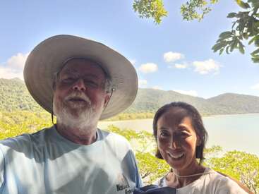 A smiling couple stands outdoors in front of a scenic lake and green mountains, with a bright blue sky and fluffy clouds above, enjoying nature together.