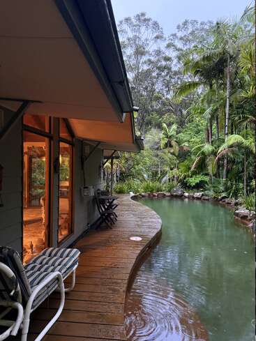A cozy house with large windows overlooks a serene, natural pool surrounded by lush tropical trees and plants. Wooden deck with seating enhances tranquility and relaxation.