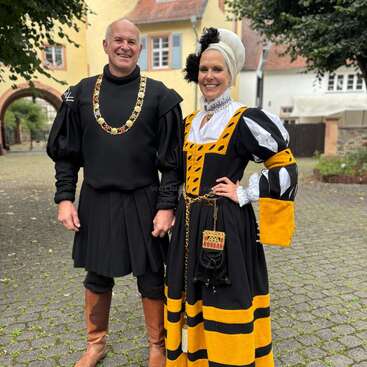 Ein Mann und eine Frau lächeln im Freien und tragen aufwendige historische Kostüme. Der Mann trägt Schwarz mit einer Kettenhalskette, die Frau ein schwarz-gelb-weißes Gewand.