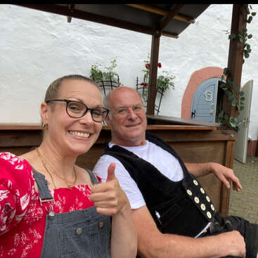 A smiling woman in glasses gives a thumbs up beside a relaxed man sitting, both in casual attire, outside near a rustic wooden structure and white wall.