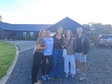 Five friends stand closely together, smiling outside on a gravel driveway with a modern house, green lawn, blue sky, and parked cars visible in the background.