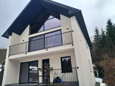 Maison moderne à deux étages avec de grandes fenêtres, un balcon et un toit en pente. Entourée d'arbres et d'un ciel couvert, l'extérieur est blanc avec des accents et des garde-corps noirs.