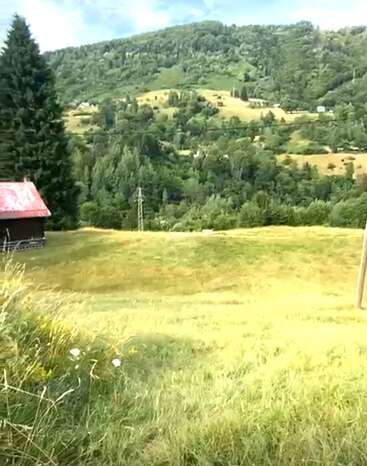 Uma paisagem rural serena apresenta uma colina gramada, uma pequena cabana de madeira, flores silvestres, linhas de energia e montanhas densamente arborizadas sob um céu azul parcialmente nublado.