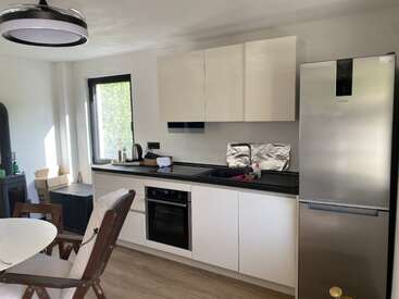 Cette image montre une cuisine moderne avec des armoires blanches, un comptoir noir, un réfrigérateur et un four en acier inoxydable, un coin repas, une fenêtre, un sol en bois et un décor minimaliste.