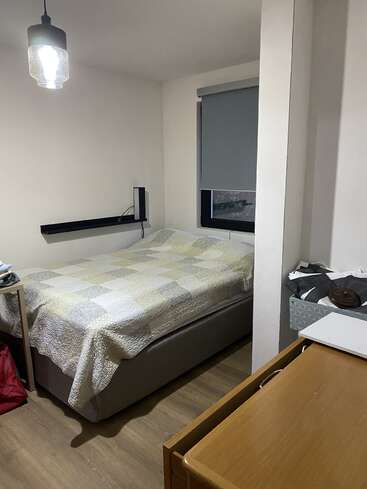 Une petite chambre bien rangée comprenant un lit avec un édredon en patchwork, un bureau en bois, un panier à linge, une fenêtre avec un store et un plafonnier.