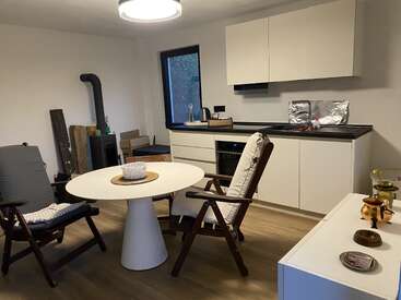 Cette cuisine confortable comprend des armoires blanches modernes, une table ronde avec des chaises rembourrées, un plancher en bois, une cuisinière à bois et divers ustensiles sur les comptoirs et le buffet.