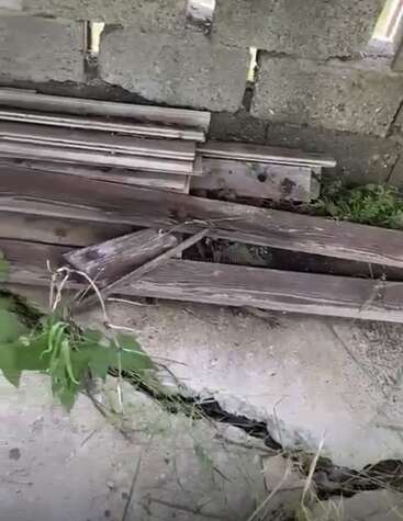 Esta imagem mostra uma superfície de concreto rachada com ervas daninhas crescidas, uma pilha de tábuas de madeira velhas e uma parede de tijolos cinza áspera ao fundo.