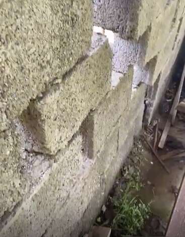 Uma visão de perto de uma parede de blocos de concreto áspera e inacabada. Alguns blocos estão desalinhados ou faltando. A vegetação e os detritos são visíveis na base do solo.