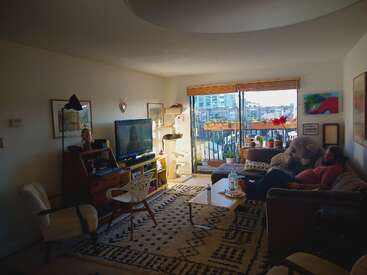 La imagen muestra una acogedora sala de estar con balcón, en la que hay un televisor, un sofá, una mesa de centro y varios muebles, con una persona descansando en el sofá.