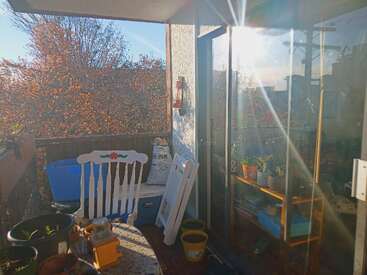 La imagen representa un acogedor balcón con una mesa, sillas y macetas, bañado por la luz del sol, rodeado de una serena vista de árboles y un cielo azul despejado.