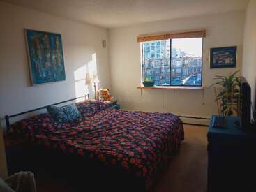 La imagen muestra un acogedor dormitorio con cama, mesilla de noche, ventana y obras de arte, con un vibrante edredón de flores y un paisaje urbano al otro lado de la ventana.