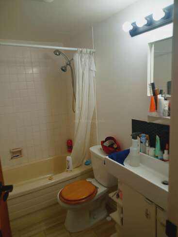 La imagen muestra un cuarto de baño con bañera, inodoro, lavabo y espejo, con azulejos blancos y una cortina de ducha blanca, y varios artículos de aseo sobre la encimera.