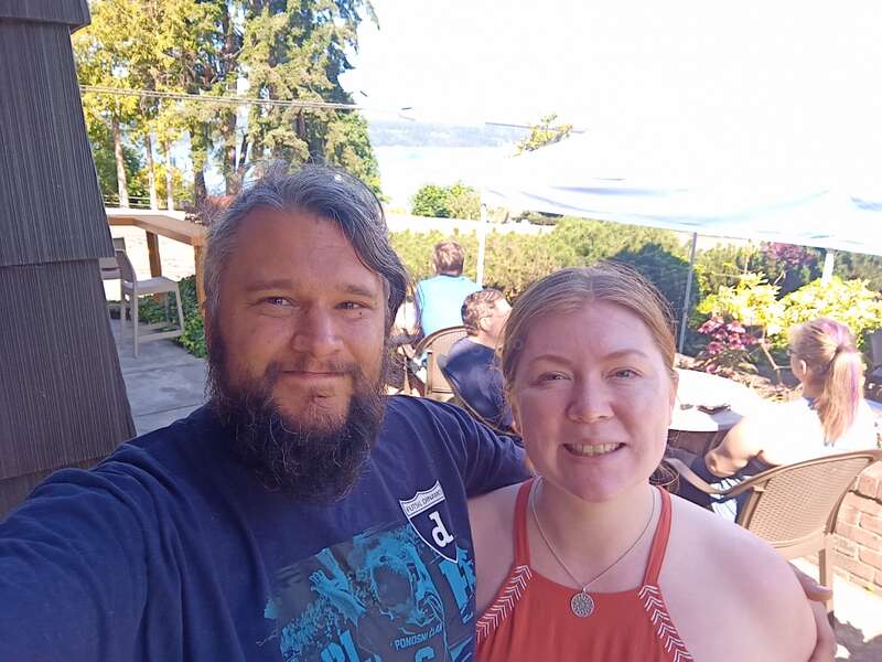 La imagen muestra a un hombre y una mujer posando para un selfie en un patio al aire libre con vistas panorámicas, rodeados de árboles y una masa de agua al fondo.