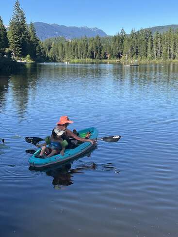 Eine Frau mit Hut und ein Kind in einer Schwimmweste paddeln mit dem Kajak auf einem ruhigen See, umgeben von Kiefern und fernen Bergen unter strahlend blauem Himmel.