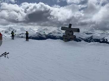 Skifahrer machen auf dem Gipfel eines schneebedeckten Berges unter dramatischen Wolken eine Pause. Ein großer Inukshuk aus Stein ragt markant empor und überblickt atemberaubende schneebedeckte Gipfel und ferne Gebirgsketten.