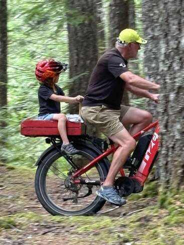 Ein Mann und ein Kind fahren mit einem roten Fahrrad durch den Wald. Das Kind trägt einen Helm und sitzt hinten auf dem Fahrrad; gemeinsam genießen sie das Abenteuer.