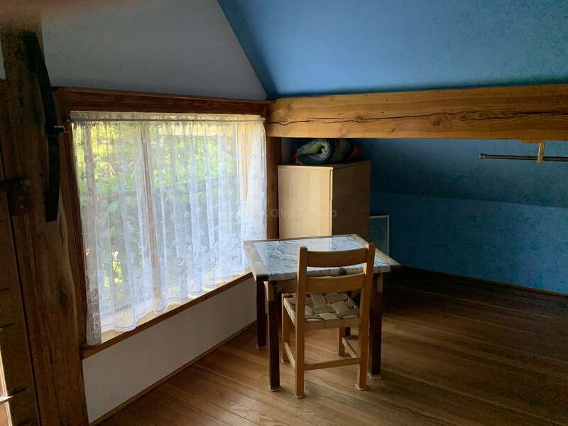 Ein kleines, gemütliches Zimmer mit Holzboden, blauen Wänden, einem Fenster mit Spitzenvorhängen, einem schlichten Stuhl, einem Tisch mit Marmorplatte, einer Kommode und bunten Kissen. Friedliche Atmosphäre.