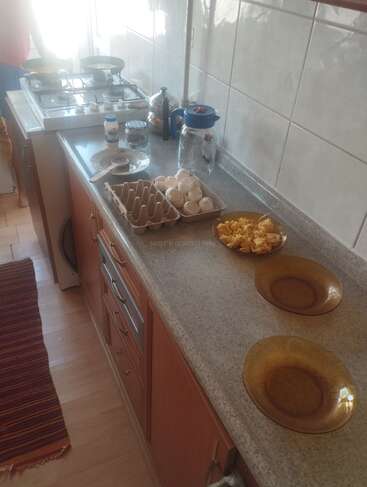 Uma bancada de cozinha com pratos marrons, ovos mexidos, cascas de ovos e utensílios de cozinha. Fogão com frigideiras, recipientes de temperos e uma jarra de vidro para água. Iluminação clara.