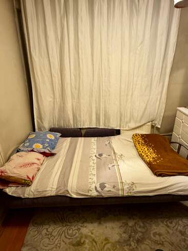 Um quarto aconchegante apresenta uma cama com lençóis de animais de desenho animado, travesseiros coloridos e um cobertor marrom, com grandes cortinas brancas e um tapete estampado.