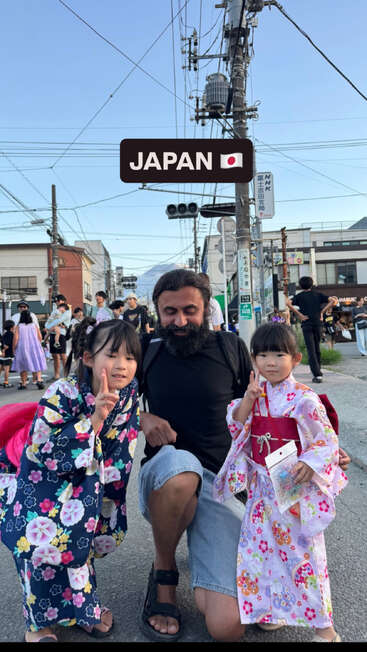 Ein Mann kniet zwischen zwei jungen Mädchen, die bunte traditionelle japanische Kimonos tragen; alle lächeln und zeigen das Peace-Zeichen auf einem Straßenfest in Japan.