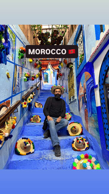 Ein Mann sitzt auf einer leuchtend blauen Treppe in Marokko, umgeben von bunten Hüten und fröhlichen Dekorationen, vor dem Hintergrund lebhafter, mit Wandmalereien verzierter Wände und hängender Blumenkörbe.