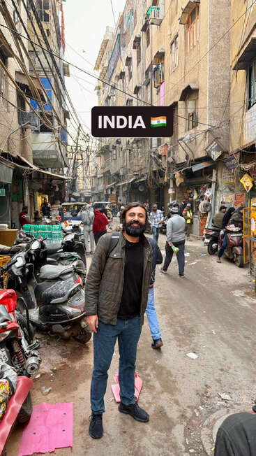 Ein Mann steht lächelnd in einer belebten, engen Straße in Indien. Motorräder und Menschen füllen das Bild, umgeben von hohen, dicht bebauten Wohnhäusern und einem Gewirr aus Kabeln.
