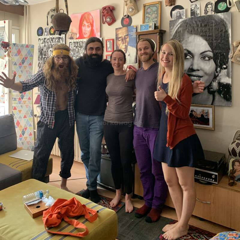 A imagem mostra um grupo de cinco pessoas em uma sala de estar, cercadas por obras de arte e decoração ecléticas. A sala está cheia de vários itens.