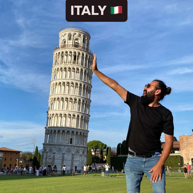 Ein Mann posiert humorvoll, als würde er den Schiefen Turm von Pisa in Italien „stützen“. Der Himmel ist blau. Touristen und grünes Gras umgeben das berühmte Wahrzeichen.