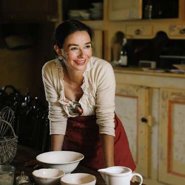 A cheerful woman with dark hair smiles warmly in a cozy kitchen, wearing a red apron and cream cardigan, surrounded by white bowls and vintage decor.