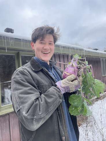 A smiling person wearing a dark jacket and gloves stands outside in snowy weather, proudly holding a large purple kohlrabi with green leaves and roots.